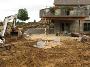 During Construction: The septic tank was exposed as a part of the excavation activities to reveal the subgrade. The two middle deck posts have been removed and a structural beam introduced to enlarge the usable lower patio space. The curved segmental block retaining walls are under construction. The aggregate base for the pavers has been thoroughly compacted to provide a solid paver base to ensure a long pavement life.