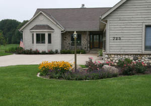 The improved landscape design provides significant curb appeal. The first step was to remove the existing Pine tree to improve visibility to the front door. In the foreground, a planting bed with a light pole, vibrant plantings and ornamental boulders extends into the yard to reduce the impact of the large concrete driveway from the street view. The planting bed is edged with lannon edger stone that lawn mower tires can drive on thereby reducing grass trimming.